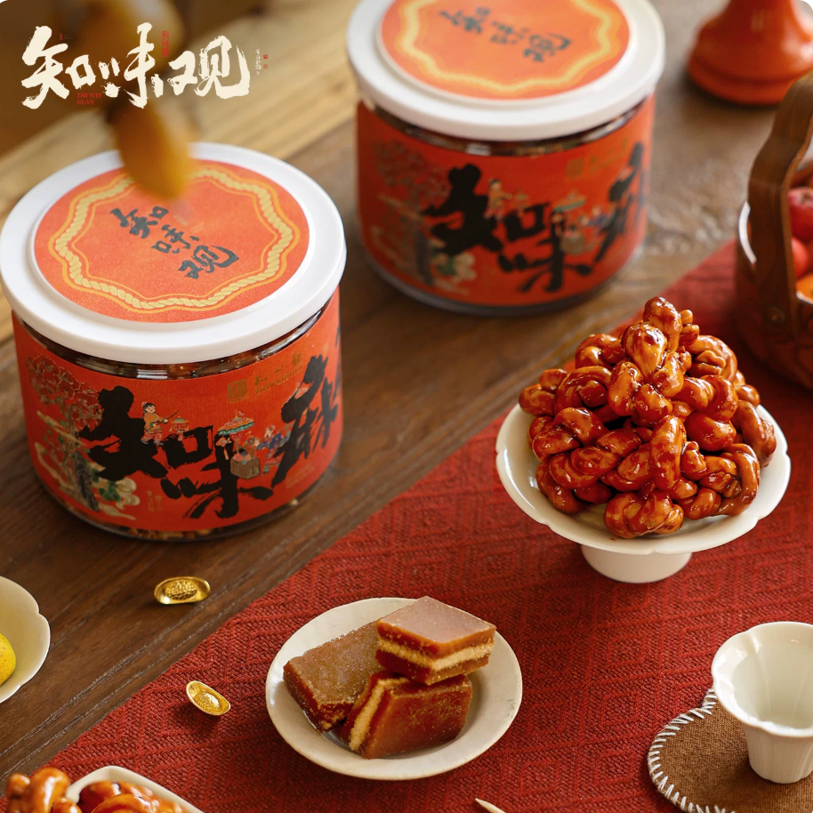 Zhiweiguan mahua two orange tins with crispy fried dough twists piled on white cake stand, festive Chinese New Year table setting
